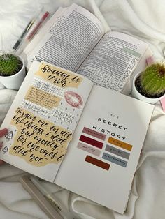 an open book on top of a bed next to two small cactuses and a pen