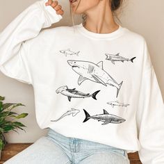 Introducing our captivating Shark Illustration Sweatshirt, a must-have for shark enthusiasts and lovers of unique apparel. This sweatshirt boasts a stunning design featuring a multitude of sharks, all intricately outlined in bold black on the front. Shark Clothing Aesthetic, White Casual Tops With Shark Design, Casual White Tops With Shark Design, Casual White Shark Design Tops, White Crew Neck Top With Shark Design, Casual Long Sleeve Sweatshirt With Shark Design, Shark Sweatshirt, Summer T-shirt With Shark Design And Short Sleeves, Casual Cotton T-shirt With Shark Design