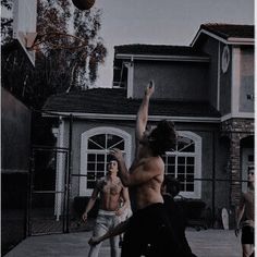 two men are playing basketball outside in front of a house with no shirt on and one man is jumping up to catch the ball