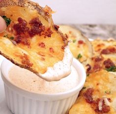 a hand holding up a piece of pizza with cheese and bacon on it, over a cup of dip