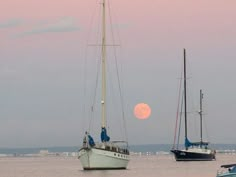 two sailboats floating in the water near each other under a pink sky at sunset