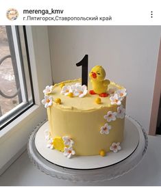 a yellow cake decorated with white flowers and a chicky on top is sitting in front of a window