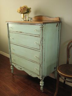 an old blue dresser with flowers on top and a chair in the corner next to it