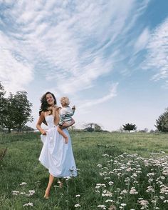 a woman in a white dress is holding a baby while standing in a field full of dandelions