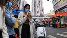 a woman wearing a face mask looking at her cell phone while standing next to a scooter