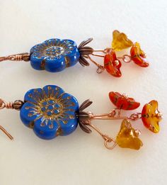 "Colorful Czech glass flower beads in cornflower blue with orange and topaz flower dangles on handmade copper ear wires. Cornflower blue Czech glass wild rose is 17mm with gold/copper Picasso etching accented with copper bead caps. Darling little mandarin orange/gold cup flowers and topaz bellflowers dangle on handmade bright copper stems. Rustic flower dangles hang on handmade hammered German copper ear wires. Total drop is 2 1/2\"." Blue Earrings With Colorful Czech Glass Beads, Picasso Flowers, Adjustable Blue Flower Earrings With Czech Glass, Blue Czech Glass Beaded Earrings, Blue Czech Glass Flower Earrings, Flower-shaped Beaded Czech Glass Earrings, Flower-shaped Jewelry With Colorful Czech Glass Beads, Blue Flower Earrings, Rustic Flower