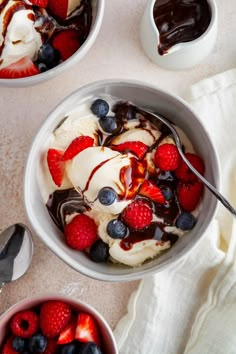 two bowls filled with ice cream, berries and chocolate sauce on top of a table