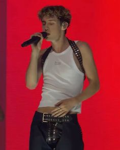 a man in white shirt and black pants holding a microphone to his mouth while standing next to a red wall