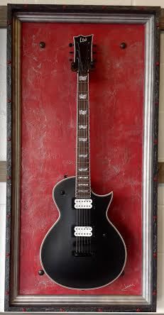 a black guitar hanging in a frame on the wall next to a red wallpaper