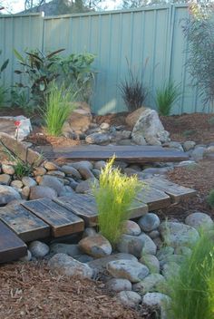 a garden with rocks and plants next to a wooden walkway that leads up to a fence