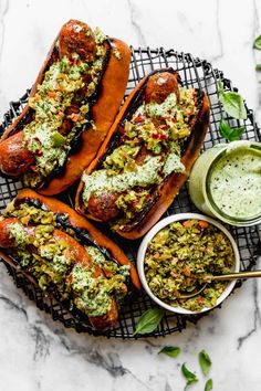 three hot dogs with toppings on a cooling rack next to dipping sauce and green herbs