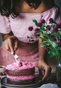 a woman is cutting into a cake with pink frosting and flowers in the background