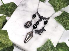 three necklaces with black beads and silver charms on a marble slab surrounded by green leaves