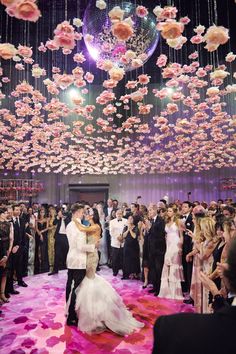 a bride and groom are kissing in front of a large group of people with pink petals floating from the ceiling