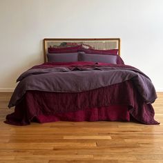 a bed with purple sheets and pillows on top of wooden floor next to white wall