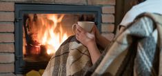 a woman wrapped in a blanket is holding a coffee cup and looking at the fireplace