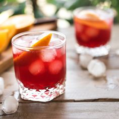 two glasses filled with red drinks sitting on top of a wooden table next to orange slices