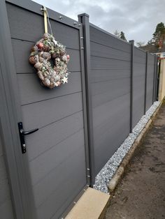 a wreath is hanging on the side of a gray fence next to a graveled area