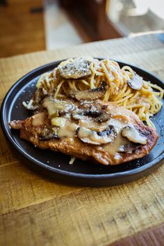 a black plate topped with meat covered in mushroom sauce and pasta on top of a wooden table