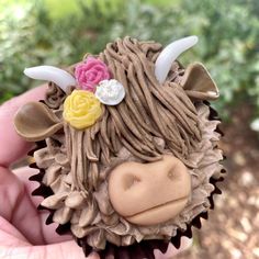 a close up of a person holding a cupcake shaped like a cow with flowers on its head