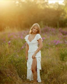 a woman in a white dress standing in a field with wildflowers at sunset