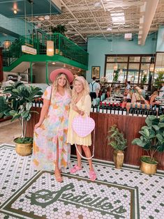 two women standing next to each other in front of a bar with plants and potted plants