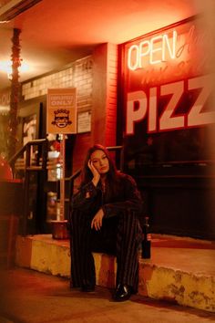 a woman is sitting on the curb in front of a pizz restaurant at night