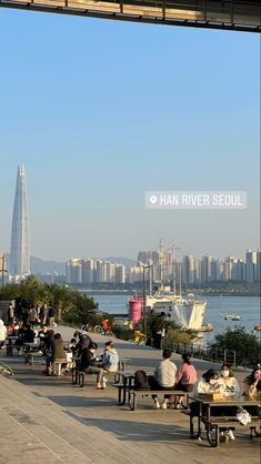 many people are sitting on benches near the water in front of a cityscape