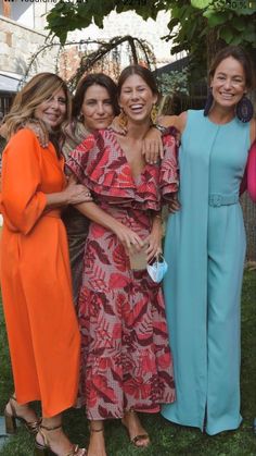 three women standing next to each other in front of a tree and grass area with one woman wearing an orange dress