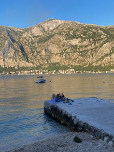 two people are laying on the edge of a pier in front of mountains and water