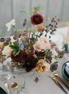 an arrangement of flowers on a table with silverware and place settings in the background