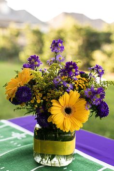 a vase filled with yellow and purple flowers on top of a green cloth covered table