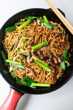 a wok filled with noodles, mushrooms and green onions next to chopsticks