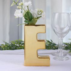 a gold letter shaped vase with flowers in it next to a wine glass on a table