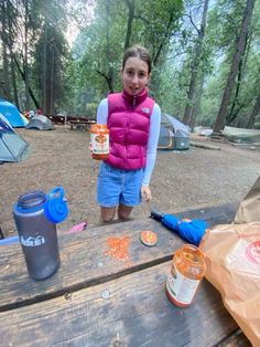 a woman standing in front of a picnic table with camping supplies on it and tents behind her