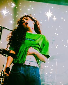 a woman standing in front of a stage wearing a green shirt and blue jeans with her arms crossed