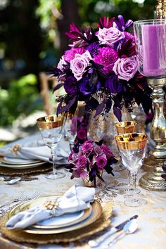 the table is set with purple flowers and gold place settings, silver candlesticks