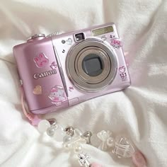 a pink camera sitting on top of a white bed next to a key chain and bracelet