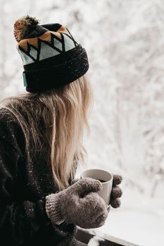 a woman holding a cup in her hand and looking out the window at snow covered trees