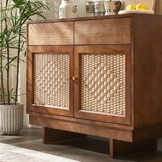 a wooden cabinet with wicker doors and handles on the bottom shelf next to a potted plant