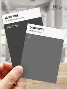 The image compares two dark gray paint swatches. On the left is "Iron Ore" by Sherwin Williams with the code SW 7069, a deep charcoal with warm undertones. On the right is "Downpipe" by Farrow & Ball with the code 26, a rich, dark gray with a slight blue-green undertone. A hand is holding both swatches, and the background is blurred with neutral interior elements. These colors are perfect for adding depth and sophistication to any space. Cozy Den, Modern Vibe