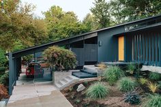 a black building with yellow door and steps leading up to the front entrance is surrounded by greenery