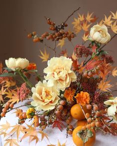 an arrangement of flowers and oranges on a white table cloth with autumn leaves in the background