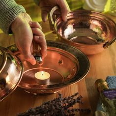 a person holding a lit candle in front of two copper dishes on a wooden table