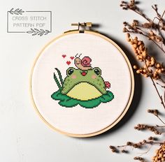 a cross stitch frog is sitting on top of a green toad with a red flower in it's mouth