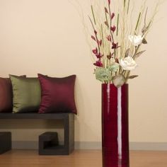 a red vase with flowers in it sitting on a wooden floor next to a bench