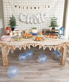 a table topped with desserts and balloons in front of a welcome to camp sign