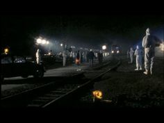 several people in white suits standing on train tracks at night with cars and lights behind them