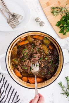 a person holding a fork in a pot filled with beef and carrots on top of a table