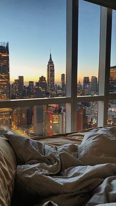 an unmade bed in front of a large window overlooking the city at night time